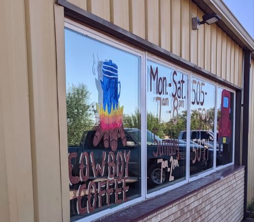 Window of a coffee shop featuring a painted coffee pot and campfire, with hours and location details.