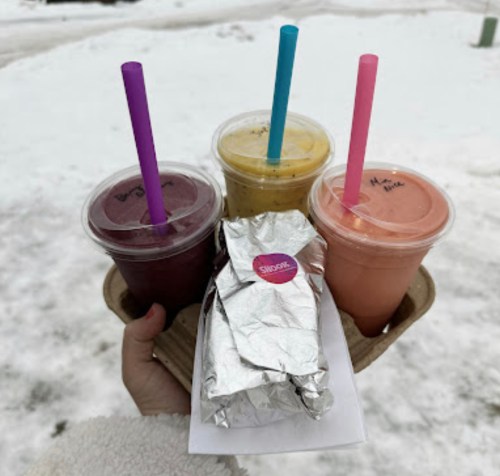 A hand holding a tray with three smoothie cups and a wrapped sandwich, set against a snowy background.
