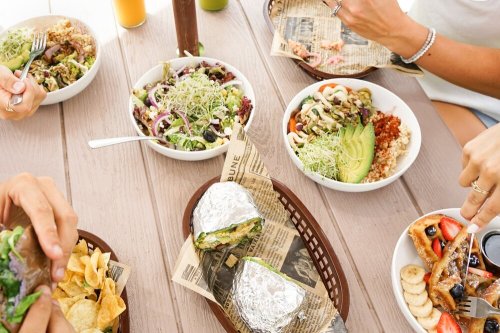 A table with various healthy bowls, a sandwich wrapped in foil, and drinks, with hands reaching for the food.