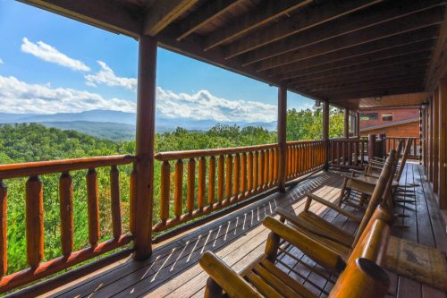 A wooden porch with rocking chairs overlooks a scenic mountain view under a blue sky with fluffy clouds.