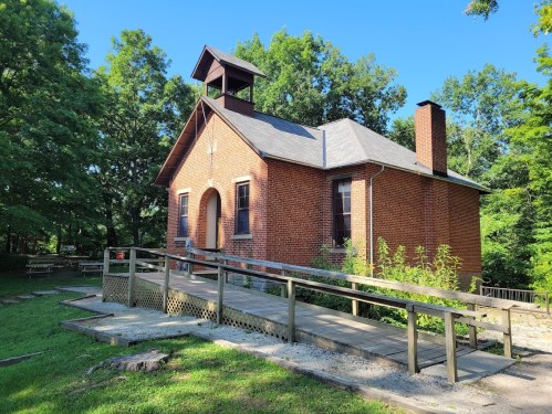 A brick building with a bell tower, surrounded by trees and a wooden ramp leading to the entrance.