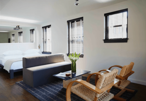 A modern bedroom with a bed, two chairs, a coffee table, and large windows with blinds.