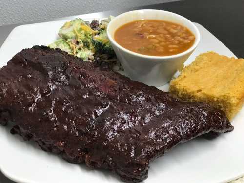 A plate featuring a large rib, cornbread, baked beans, and a side salad with broccoli and cheese.