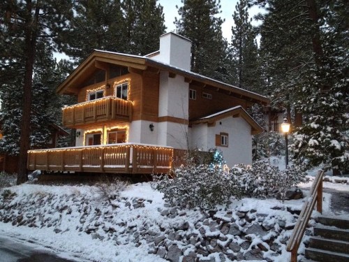 A cozy wooden house decorated with lights, surrounded by snow and tall pine trees in a winter setting.
