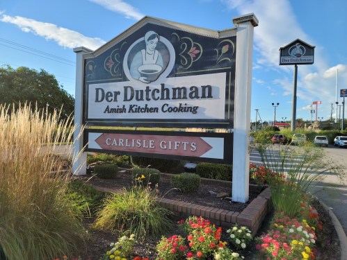 Sign for Der Dutchman, featuring "Amish Kitchen Cooking" and "Carlisle Gifts," surrounded by greenery and blue sky.