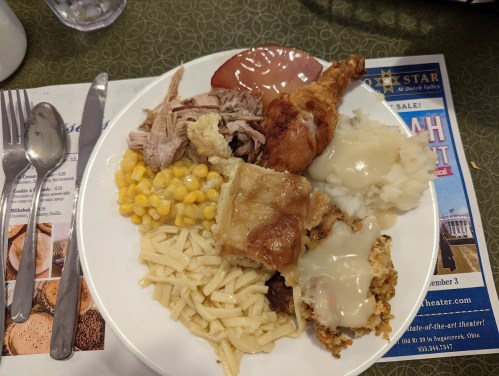 A plate of food featuring fried chicken, pulled pork, corn, mashed potatoes with gravy, noodles, and stuffing.