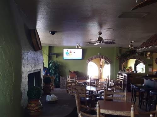 Interior of a restaurant featuring wooden tables, a bar, a TV on the wall, and decorative plants.