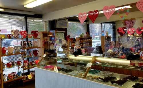 A cozy chocolate shop with heart-shaped decorations, showcasing various chocolates and treats behind a glass display.