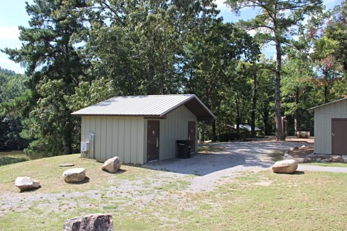 A small, single-story building with a metal roof, surrounded by trees and rocks, on a gravel path.