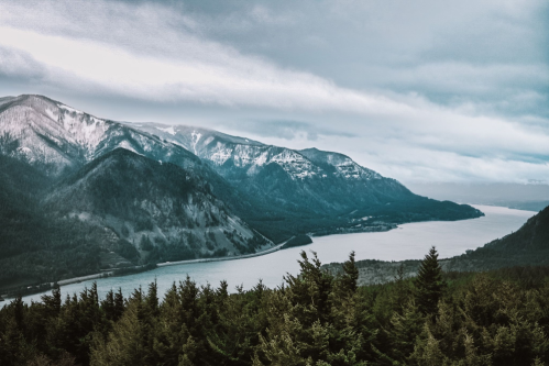 A panoramic view of a river winding through mountains, surrounded by dense forests under a cloudy sky.