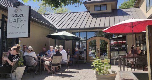 Outdoor café scene with patrons seated at tables under umbrellas, enjoying drinks and conversation on a sunny day.