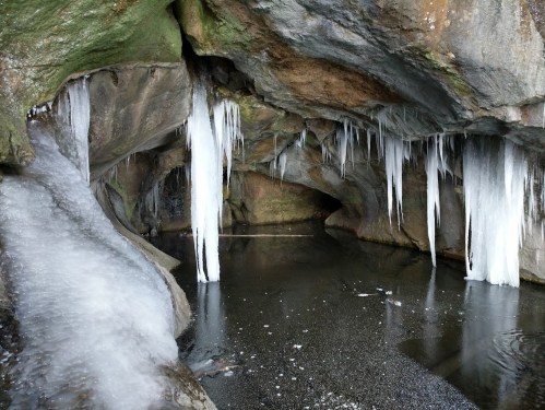 A serene cave with icicles hanging from the ceiling and a still, dark pool of water on the ground.