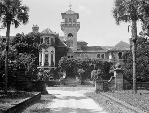A historic mansion with ivy-covered walls, a tower, and palm trees, set along a gravel driveway.