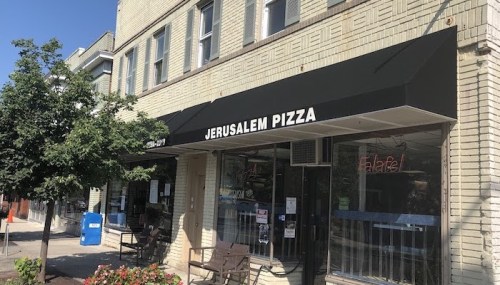 Exterior of Jerusalem Pizza restaurant with a black awning, outdoor seating, and nearby greenery.