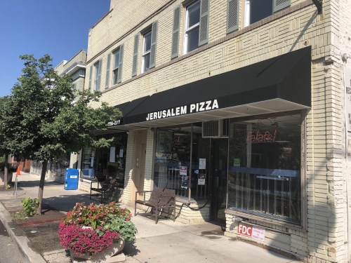 Exterior of Jerusalem Pizza restaurant with a black awning, outdoor seating, and colorful flower planters.