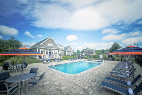A sunny pool area with lounge chairs and umbrellas, surrounded by a fence and a residential building in the background.
