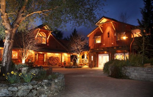 A warmly lit rustic house at night, surrounded by trees and flowers, with a stone pathway leading to the entrance.