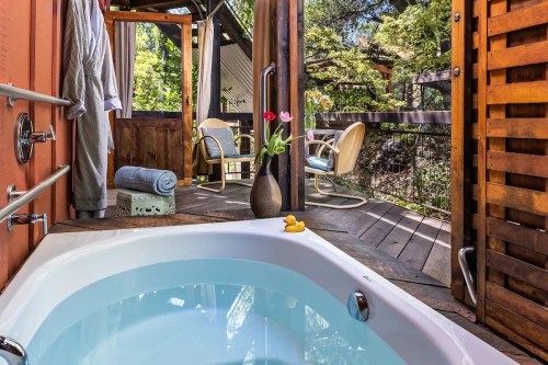 A cozy outdoor bathroom with a hot tub, wooden deck, and a view of greenery, featuring a vase of flowers and a rubber duck.