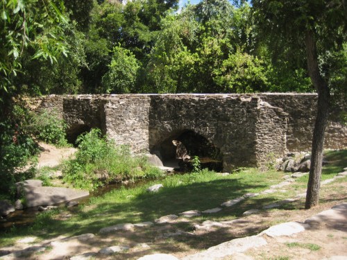 A stone bridge with arches spans a small stream, surrounded by lush greenery and trees in a sunny park setting.