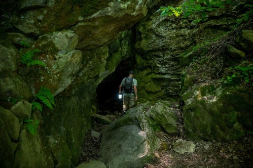 A person with a backpack and flashlight explores a rocky cave surrounded by lush greenery.