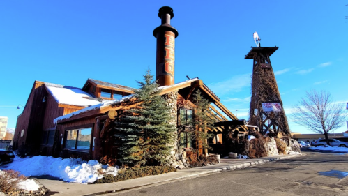 A rustic building with a tall chimney, surrounded by snow and trees, under a clear blue sky.