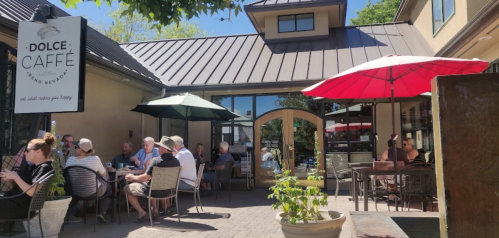 Outdoor café scene with patrons sitting at tables under umbrellas, enjoying drinks and food in a sunny setting.