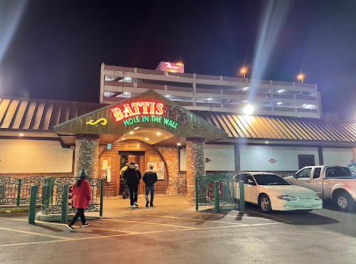 A restaurant entrance at night, with a sign reading "Battis Hole in the Wall" and a parking garage in the background.