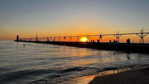 Silhouette of a pier at sunset, with people walking along the structure and reflections on the calm water.