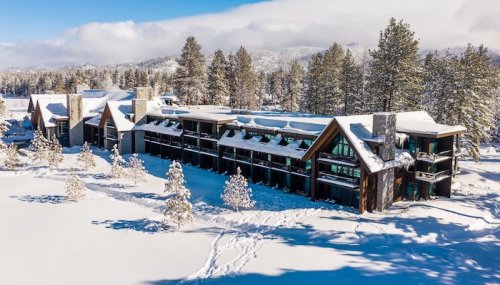 A snowy landscape featuring a modern lodge surrounded by tall trees and mountains under a clear blue sky.
