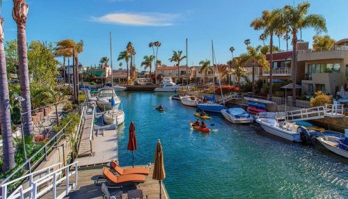 A sunny canal scene with boats and kayakers, surrounded by palm trees and waterfront homes.