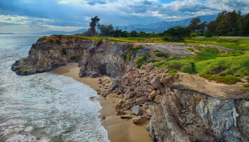 A scenic coastal view featuring rocky cliffs, sandy beach, and waves under a cloudy sky with distant mountains.