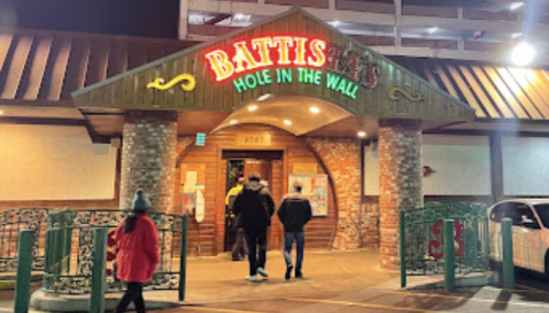 Entrance of "Battis Hole in the Wall" restaurant, with people walking in and colorful signage illuminated at night.