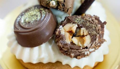 Close-up of a dessert topped with chocolate candies, nuts, and a sprinkle of gold dust on creamy frosting.
