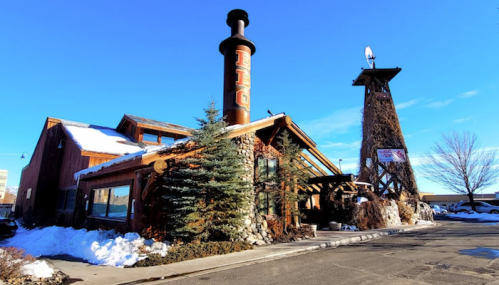 A rustic building with a tall chimney, surrounded by snow and trees, under a clear blue sky.