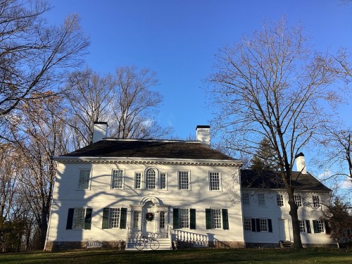 A large white historic house with green shutters, surrounded by bare trees against a clear blue sky.