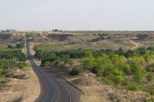 A winding road stretches through a hilly landscape with sparse trees and open fields under a clear sky.