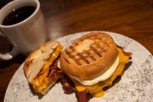 A toasted sandwich with bacon, cheese, and egg, served on a plate next to a cup of black coffee.