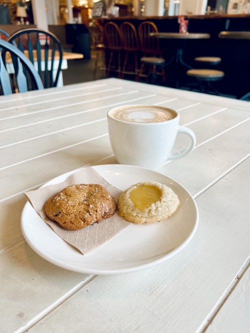 A cup of latte on a table with two cookies on a plate, one chocolate chip and one lemon.