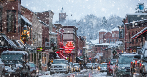 A snowy street scene with charming shops and cars, illuminated by a glowing sign for a log cabin restaurant.