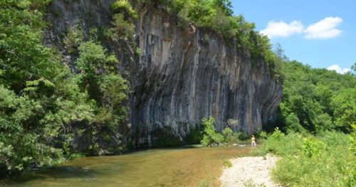 A serene river flows beside a tall, rocky cliff, surrounded by lush greenery under a clear blue sky.