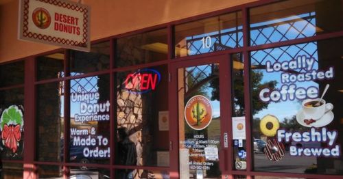 Exterior of Desert Donuts shop with "OPEN" sign, showcasing colorful window graphics for donuts and coffee.