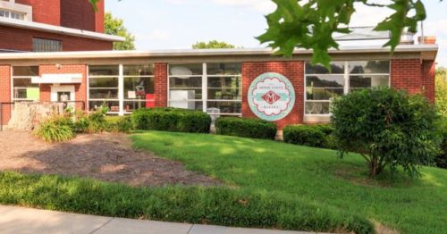 A brick building with large windows and a circular sign, surrounded by green grass and shrubs.
