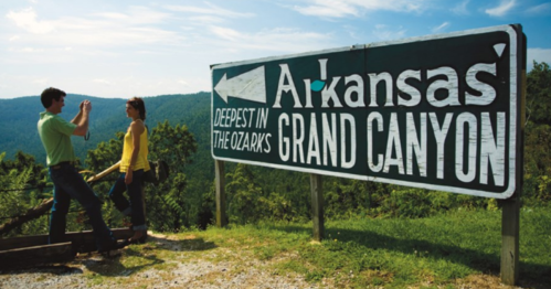 A sign reading "Arkansas' Grand Canyon" with two people taking photos in a scenic outdoor setting.