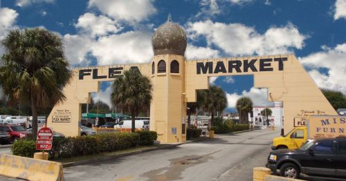 Entrance to a flea market with a large sign, palm trees, and a blue sky with clouds in the background.