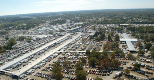 Aerial view of a large outdoor market with numerous stalls and parked vehicles, surrounded by trees and open space.
