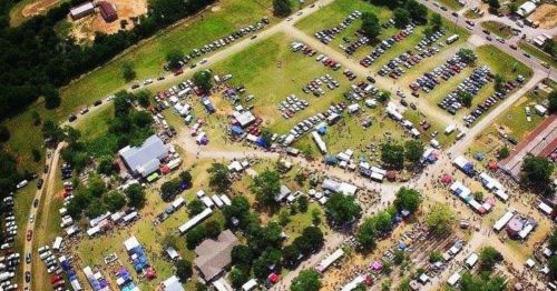 Aerial view of a large outdoor event with numerous tents, vehicles, and people gathered on green grass.