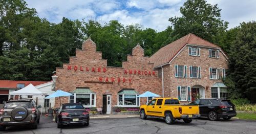 A brick building with "Holland American Bakery" sign, surrounded by trees and parked cars, under a blue sky.