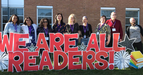 A group of diverse individuals stands in front of a large sign that reads "WE ARE ALL READERS!" with festive decorations.