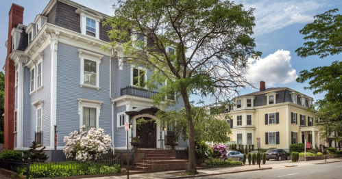 A charming blue Victorian house with white trim, surrounded by greenery and blooming flowers, on a sunny street.