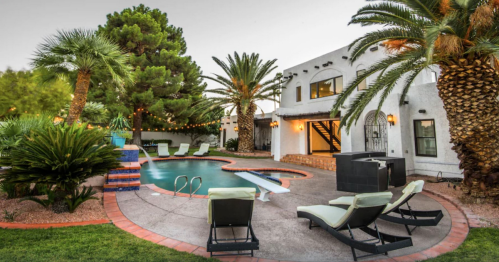 A serene backyard with a pool, palm trees, lounge chairs, and a white house, creating a relaxing outdoor space.